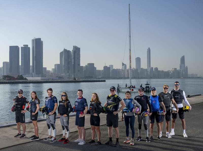 Equipe de velejadores posando à beira-mar com skyline urbano ao fundo durante evento de vela internacional