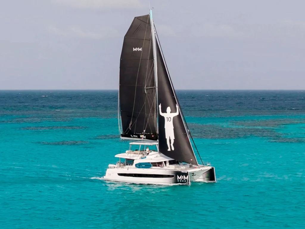 Catamarã Bali 5.4 com velas pretas personalizadas com a silhueta de Lionel Messi navegando em águas cristalinas.