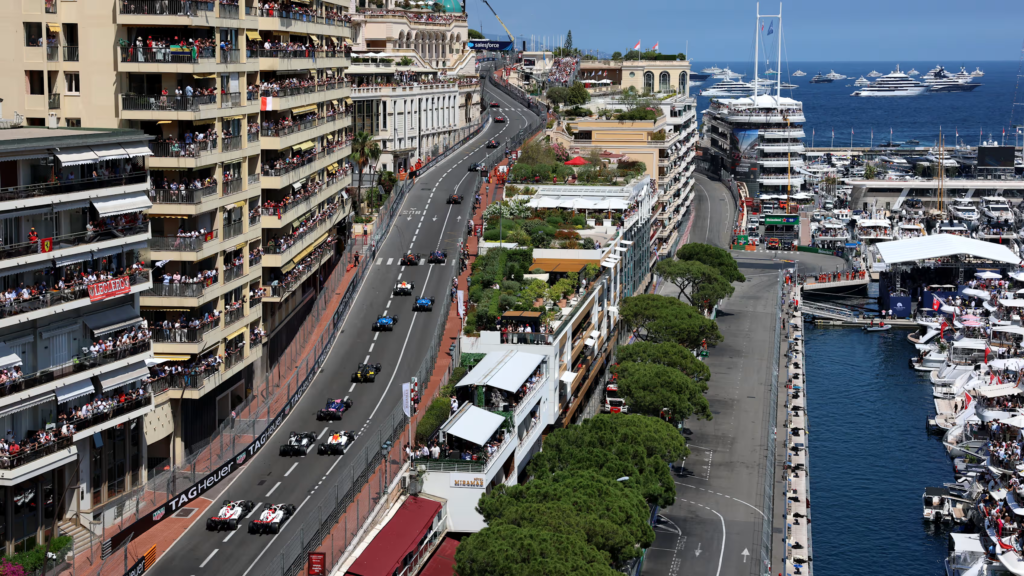 Carros de Fórmula 1 percorrem as ruas de Monte Carlo enquanto megaiates aparecem ancorados ao fundo no Mediterrâneo.