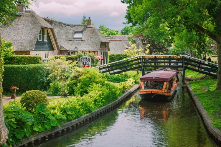 Barco navegando por canal de Giethoorn entre casas tradicionais com telhados de palha, jardins e ponte de madeira na Holanda.