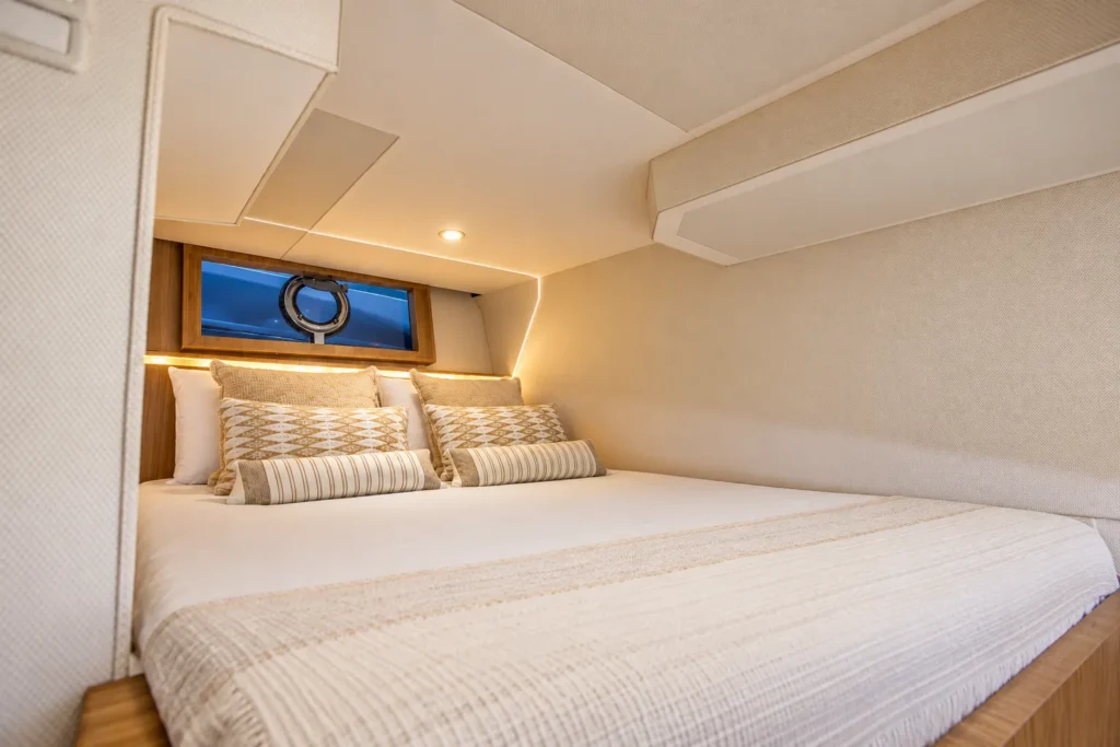 Cabine da Coral 40 Sea Breeze com cama de casal, roupa de cama clara, almofadas decorativas e janela circular na cabeceira.