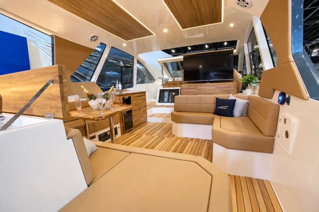Salão da Coral 40 Sea Breeze com sofás bege, piso em teca, mesa de apoio, televisão retrátil e janelas amplas.
