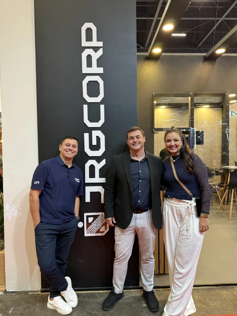 Gean Schneider, Januario Gagliardi e Carol Santiago posam juntos em frente ao painel da JRG Corp durante o São Paulo Boat Show 2026, com Januario ao centro. Os três estão sorrindo para a câmera.